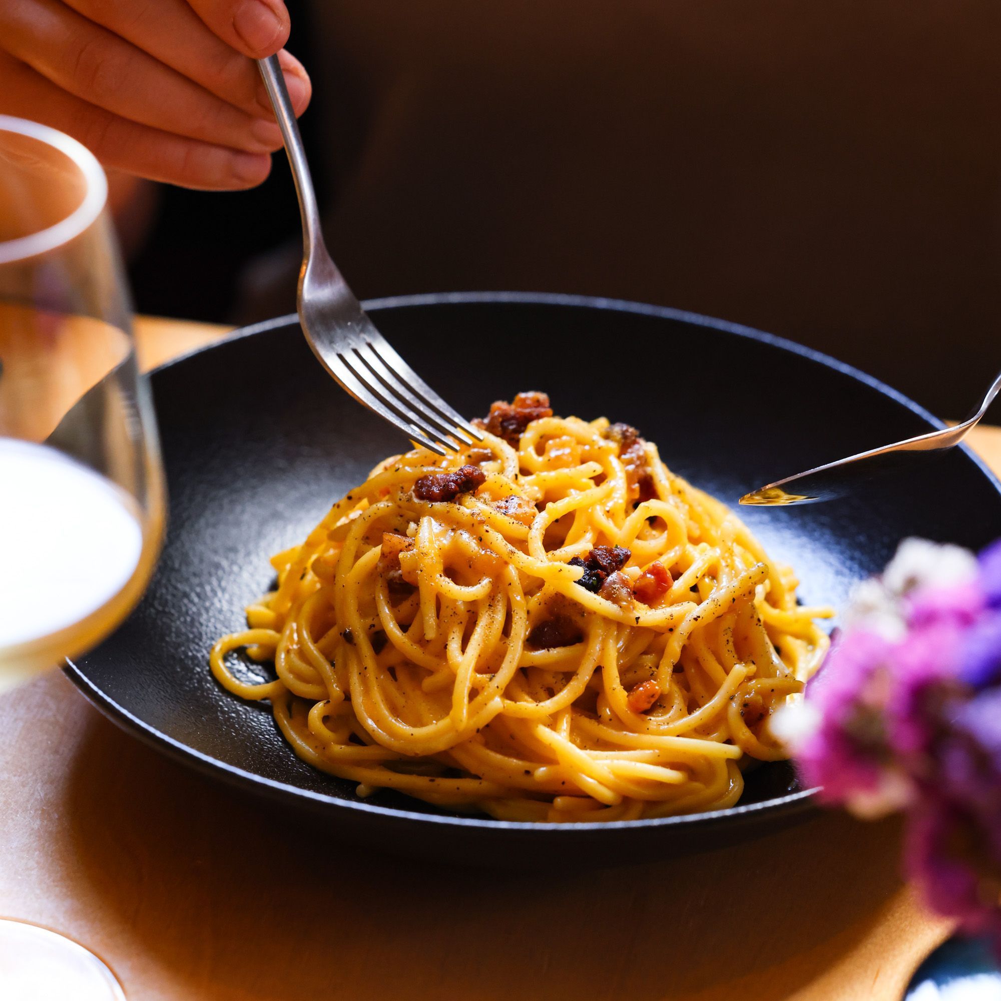 Spaghetti carbonara with crispy guanciale in a black bowl, a glass of white wine beside it